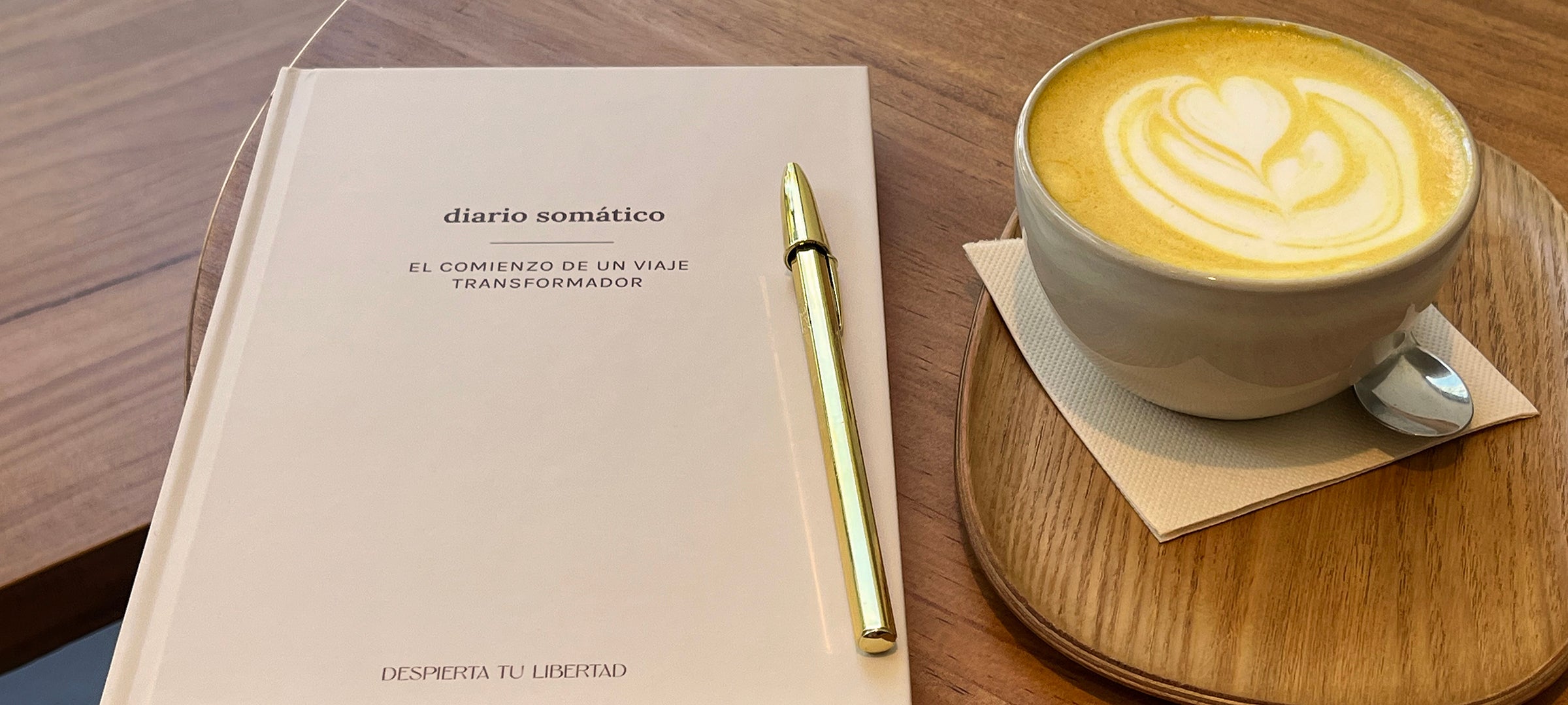 Diario somático junto a una taza de café y un bolígrafo dorado sobre una mesa de madera, invitando a la reflexión y al autoconocimiento